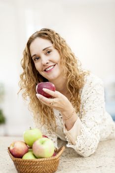Portrait of smiling woman holding red apple leaning on countertop in kitchen at home