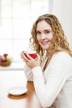 Portrait of smiling woman holding red coffee cup sitting at table in home kitchen by window
