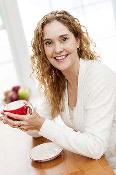 Portrait of smiling woman holding red coffee cup sitting at table in home kitchen by window