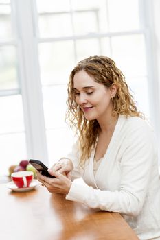 Portrait of smiling woman holding cell phone sitting at table in home kitchen by window