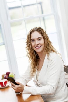 Portrait of smiling woman holding cell phone sitting at table in home kitchen by window