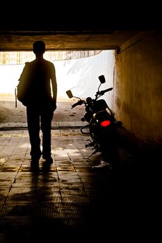 Vertical silhouette concept capture of a man in a vest with ruck sack back pack near his motorcycle which has its tail slights switched on in a tunnel