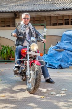 Color vertical portrait of a trendy fashion driven senior motor cyclist sat back on  a red cruiser smiling neutral expression in leather, denim, scarf and sunglasses. Generic shot location Bombay India with model release