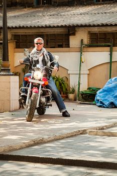 Color vertical portrait of a trendy fashion driven senior motor cyclist sat back on  a red cruiser smiling neutral expression in leather, denim, scarf and sunglasses. Generic shot location Bombay India with model release