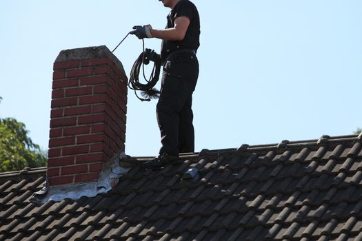 Chimney sweep cleaning a chimney standing balanced on the apex of a house roof lowering equipment down the flue