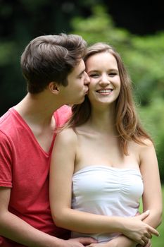 Loving young man kissing his pretty smiling teenage girlfriend on the cheek as they stand in a close embrace in a lush green garden
