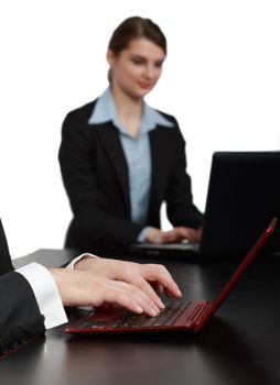 Close-up image of masculine hands working on a red notebook in front of a young woman using a laptop on a black table in an office.