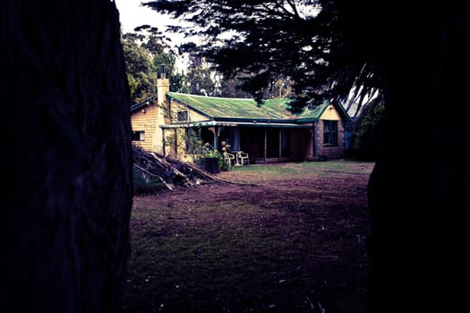 Dark toned image of an old rural farmhouse with an iron roof viewed from between two tree trunks