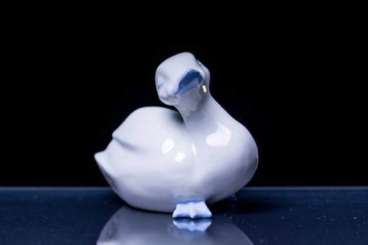 a white porcelain duck with light blue inserts standing on a reflective black surface with black background