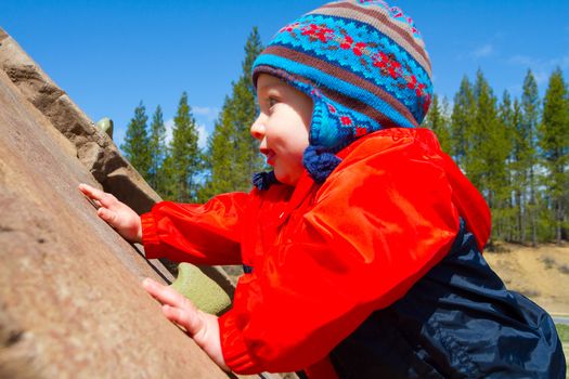 A one-year-old boy plays at a park outdoors with wood chips and structures to keep him entertained.