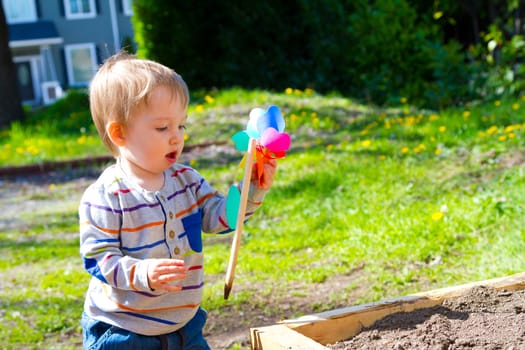 A one year old boy plays with a whirligig propeller pinwheel outside while wearing a striped shirt.