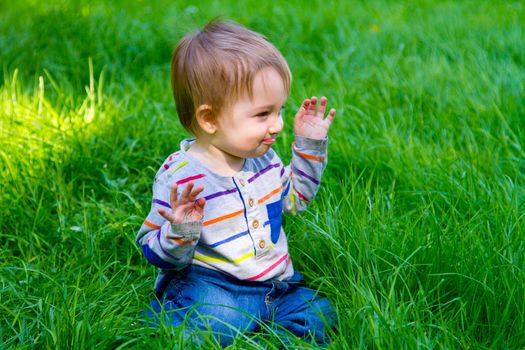 A one year old toddler boy plays in the grass in his backyard while waving and looking cute and smiling.