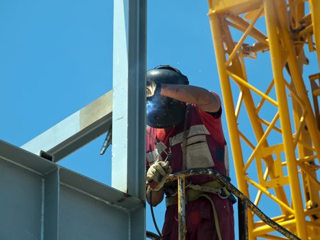 welder with protective mask welding metal construction, yellow crane on his back