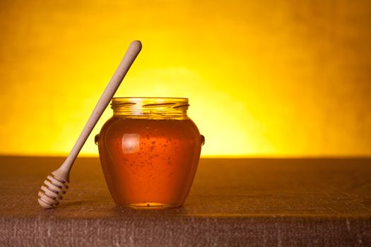 Glass honey jar with wooden dipper over yellow background   