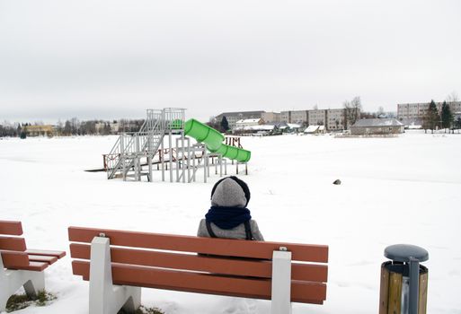 woman in grey coat sit on wooden bench in beach playground covered with snow and cascade water equipment.