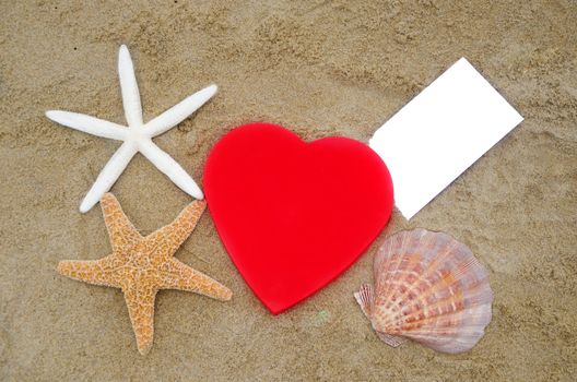 Red Heart shape, two starfishes, big seashell and piece of paper on the sandy beach