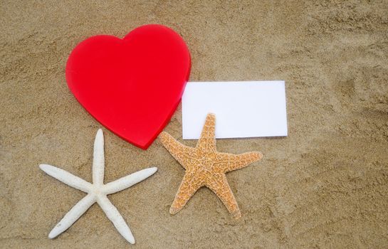 Red Heart shape, two starfishes, and piece of paper on the sandy beach