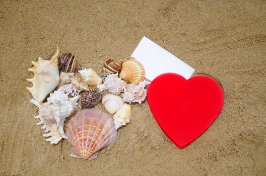 Two Heart shapes - red plastic and of seashells, with piece of paper on sandy beach