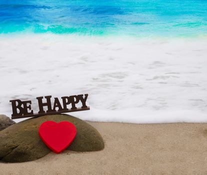 Red Heart shape and inscription "Be happy" on sandy beach by the ocean on background