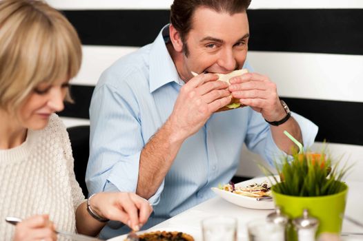 Man eating sandwich in a restaurant along with his wife.