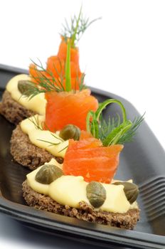 Three Snacks with Smoked Salmon, Greens, Cheese Cream and Capers on Black Plate closeup on white background