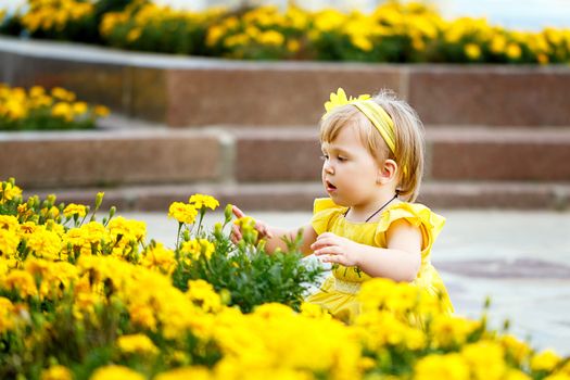 Girl in a yellow dress sits near a flower bed with yellow flowers