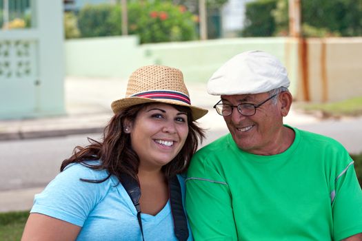 A older Hispanic senior citizen man sits outdoors in a tropical setting with his granddaughter.