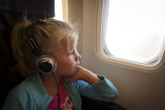 A little girl with headphones looking out of the window of an airplane