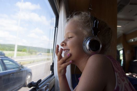 A happy girl with eyes closed sits by window in moving trailer with headphones on