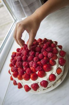 A layered cake in the kitchen, with a hand placing the final berry on top