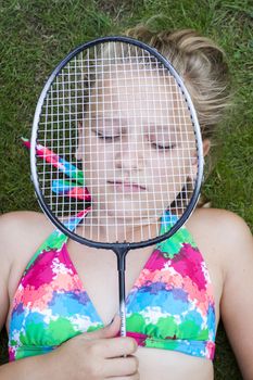A teenage girl lying on the lawn outside with eyes closed, holding a badminton racket in front of her face