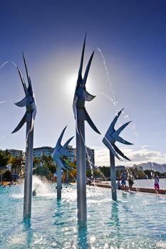 Steel fish sculpture and fountain at the Esplanade in Cairns with people playing in the water of the swimming lagoon on a hot summer day