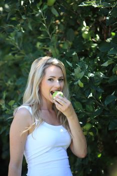 Attractive young woman eating a green apple while standing outdoors in a summer garden