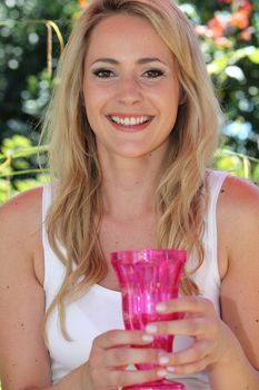 Smiling beautiful young woman holding a vibrant pink glass vase in her hands while standing outdoors on a summer day