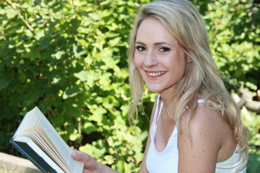 Portrait of a beautiful young blond woman reading a book outdoors in front of leafy greenery looking at the camera with a smile