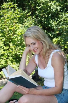 Attractive young blond woman reading a book outdoors in the garden in the shade of a tree