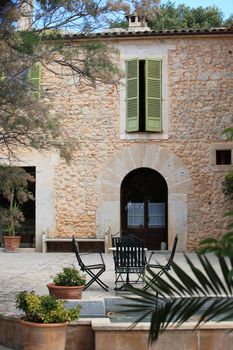 Table and chairs standing in a courtyard in front of a beautiful old stone building with an arched door and shuttered windows