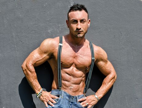 Muscular bodybuilder shirtless with jeans and suspenders against grey wall