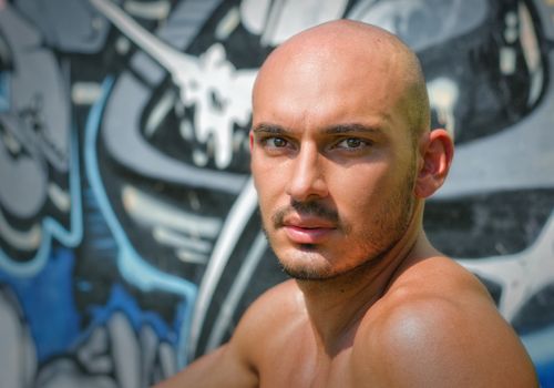 Headshot of bald young man shirtless outdoors in front of graffiti wall