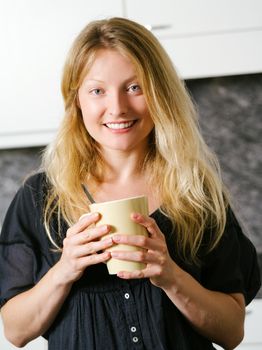 Photo of a beautiful smiling blond female in her kitchen holding a large coffee.
