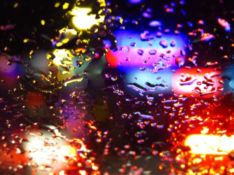 A background of a glass covered with raindrops with a backdrop of vehicle lights at night.                               