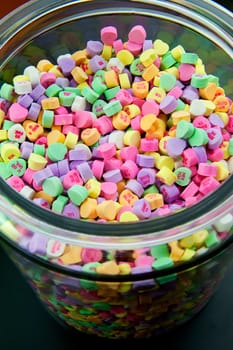 Large glass jar of colorful candy hearts with lid sits on a black table with a white background