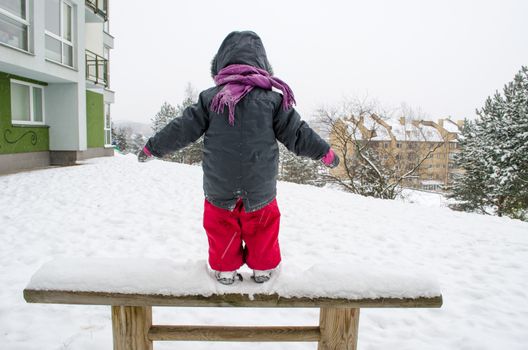 child with bright red pants and pink gloves standing on a wooden bench snow in winter time