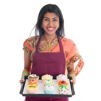 Traditional Indian woman in sari baking bread and cupcakes, wearing apron holding tray isolated on white.