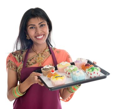 Happy Traditional Indian woman in sari baking bread and cupcakes, wearing apron holding tray isolated on white.