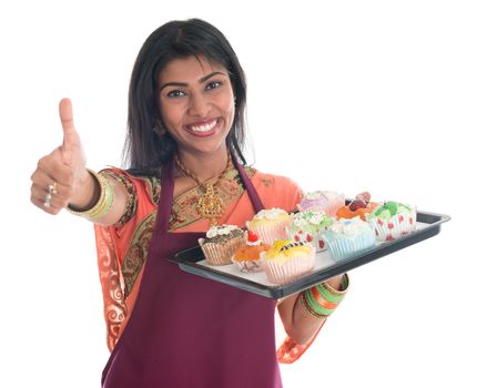 Happy Traditional Indian woman in sari baking bread and cupcakes and showing thumb up, wearing apron holding tray isolated on white.