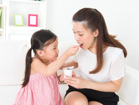 Eating yogurt. Happy Asian family eating yoghurt at home. Beautiful child feeding mother, healthcare concept.