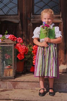 Little girl in a dirndl in front of a farmhouse with a bouquet of flowers