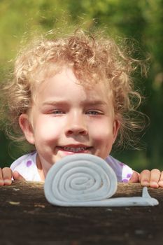 Smiling beautiful little girl with wonderful curly blond hair crouched low looking at a coiled toy snail on an outdoor wooden table