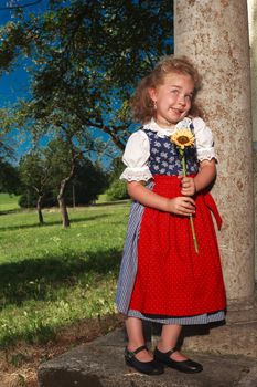 Bavarian girl with a charming little smile, leaning against a pillar with a sunflower in the hands